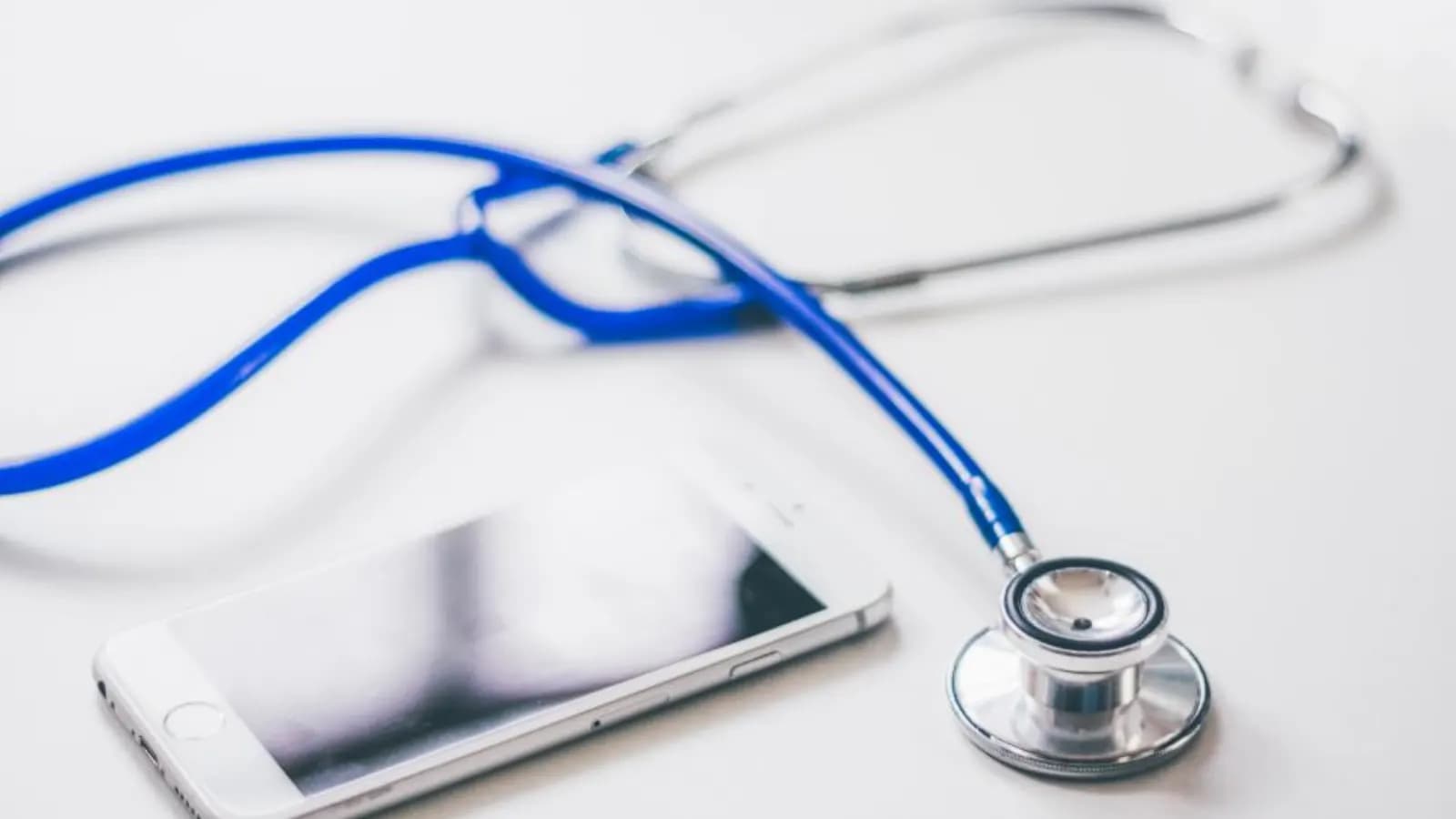 A blue stethoscope lying next to a smartphone on a white desk