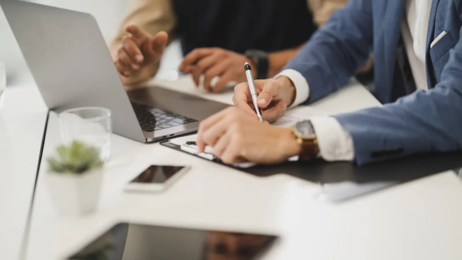 Two people at a white desk, one writing notes on a clipboard beside an open laptop