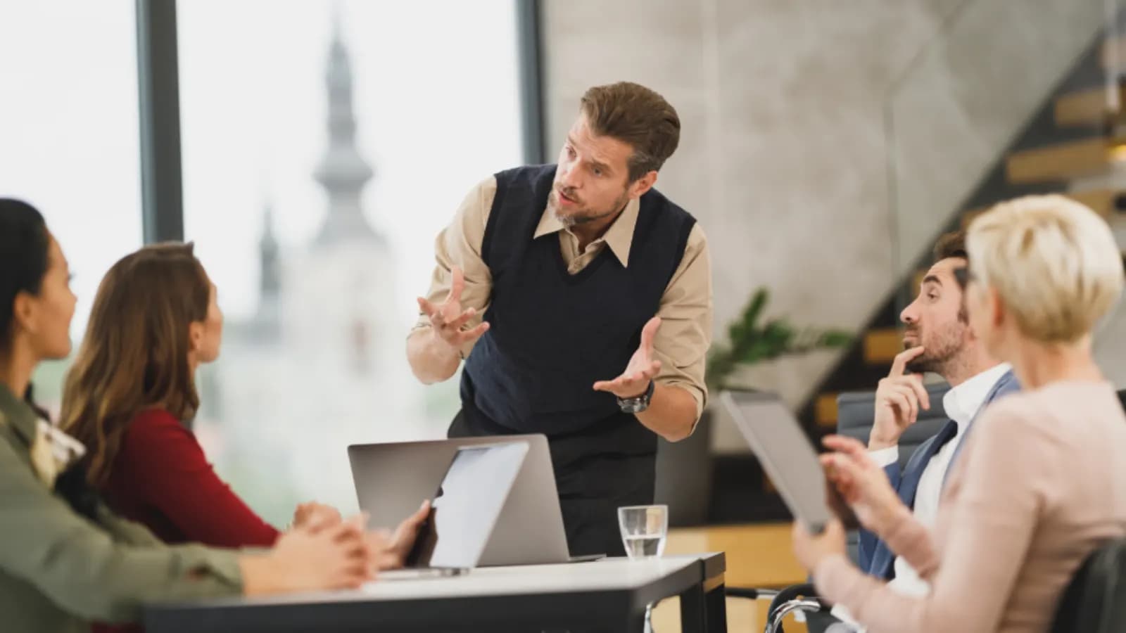 Manager standing at a meeting table explaining a plan to a team seated with laptops