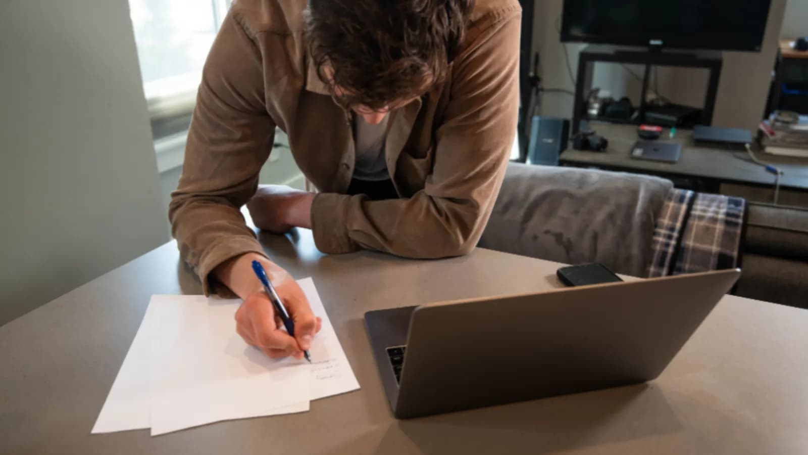 Man leaning over a table writing notes on paper next to an open laptop and a phone