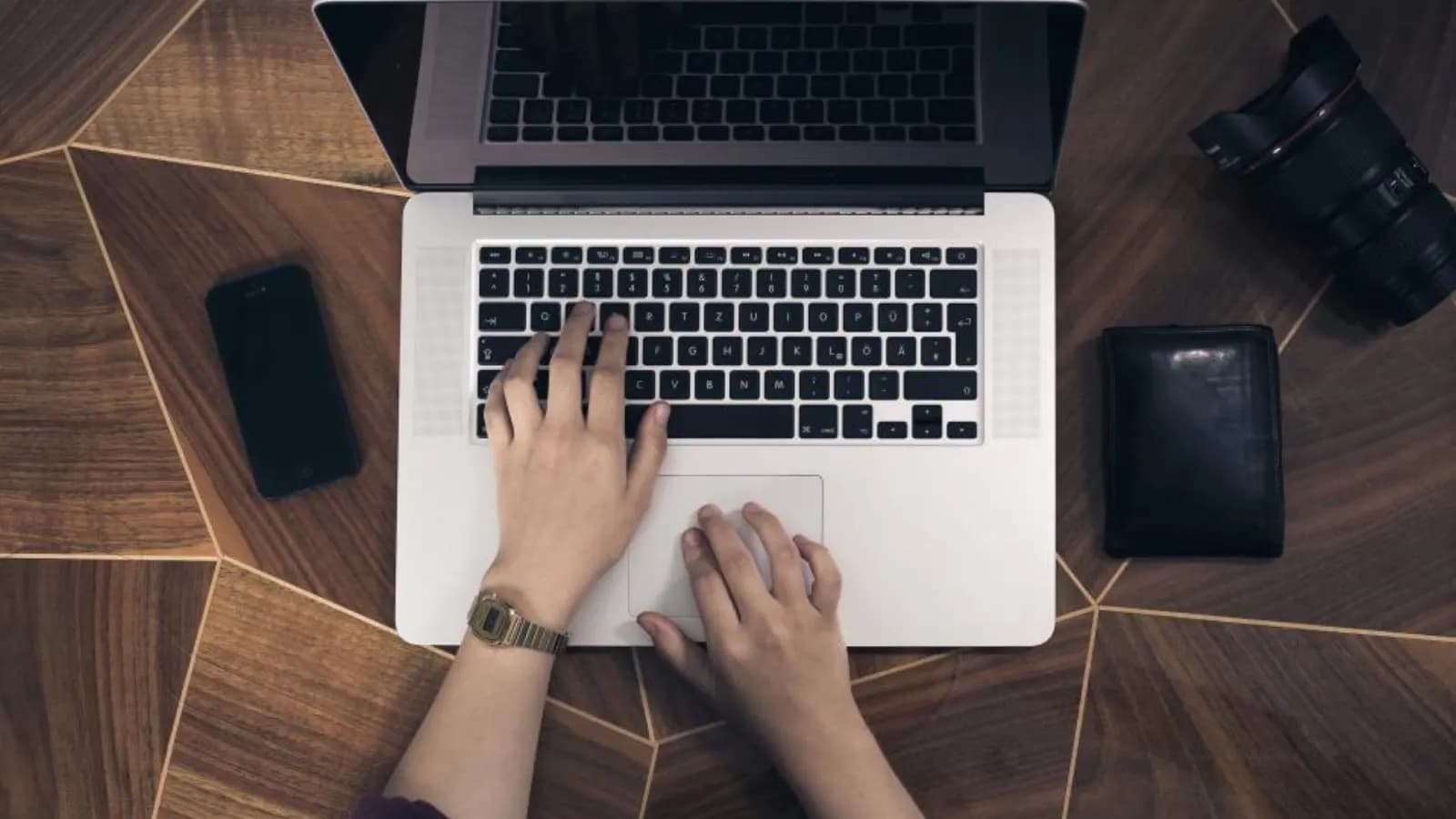 Overhead view of hands typing on a laptop on a wooden desk, with a phone and wallet nearby